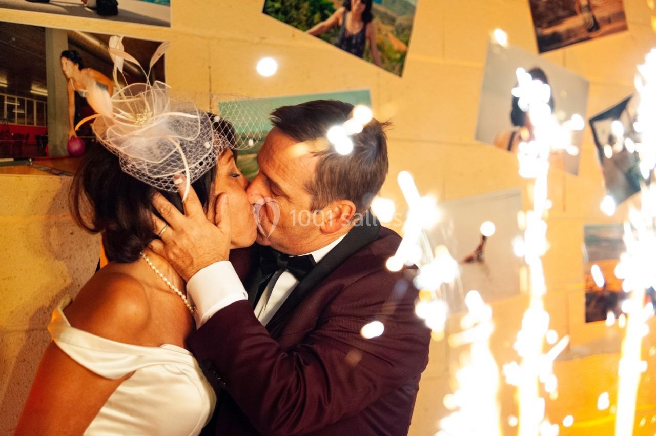 Un couple en tenue de mariage s'embrasse passionnément devant un mur décoré de photos et des étincelles.