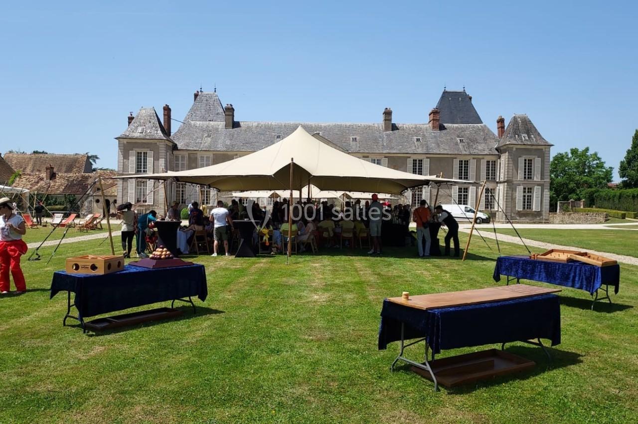 Tente dressée devant un château, entourée de tables de jeux en bois et de personnes rassemblées sur une pelouse.