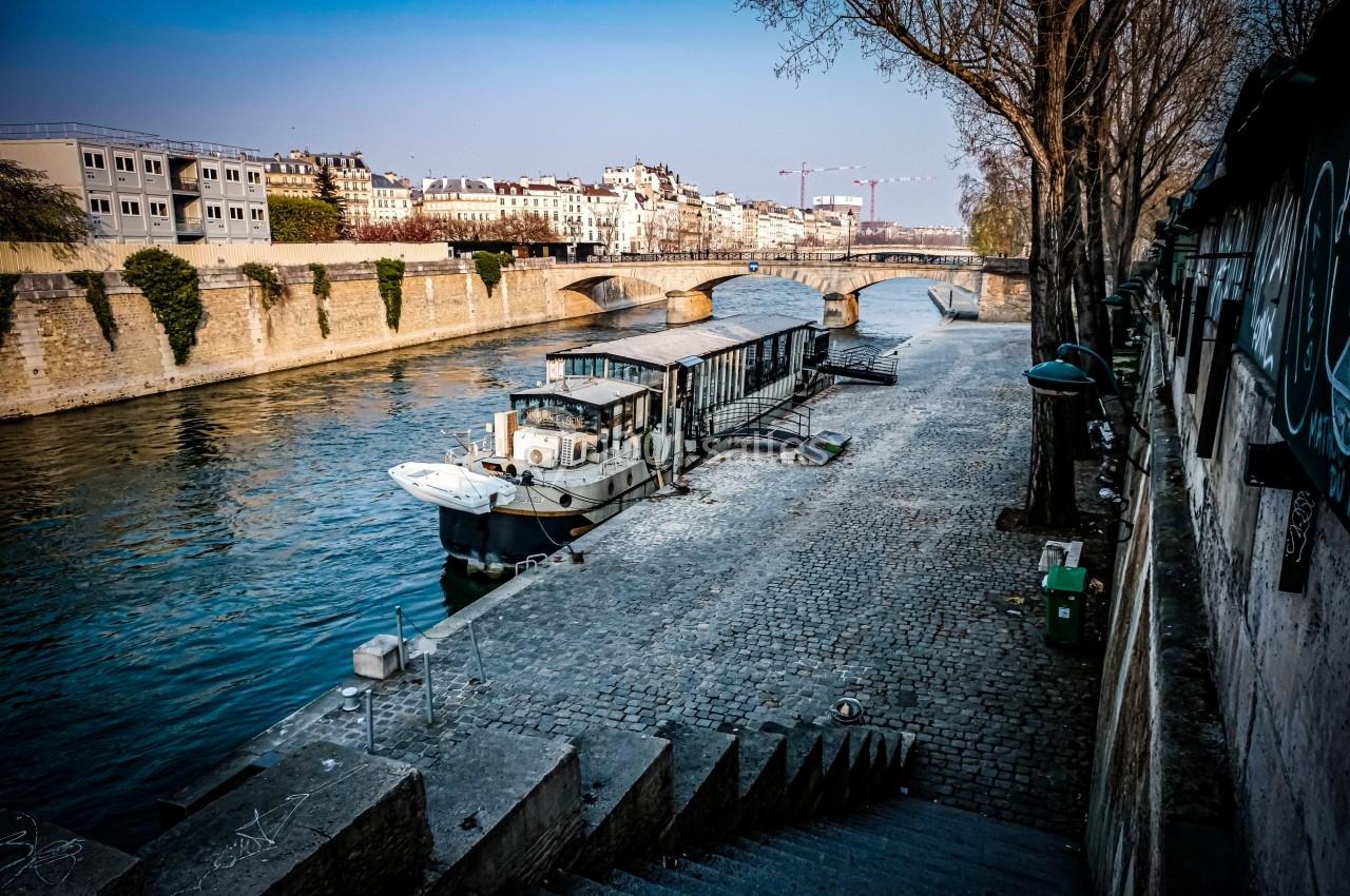 Bateau amarré sur une rivière bordée de quais pavés, avec des immeubles et un pont en arrière-plan.