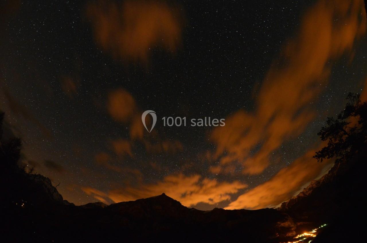 Ciel étoilé avec des nuages illuminés par des lumières orangées, montagnes et village éclairé en contrebas.