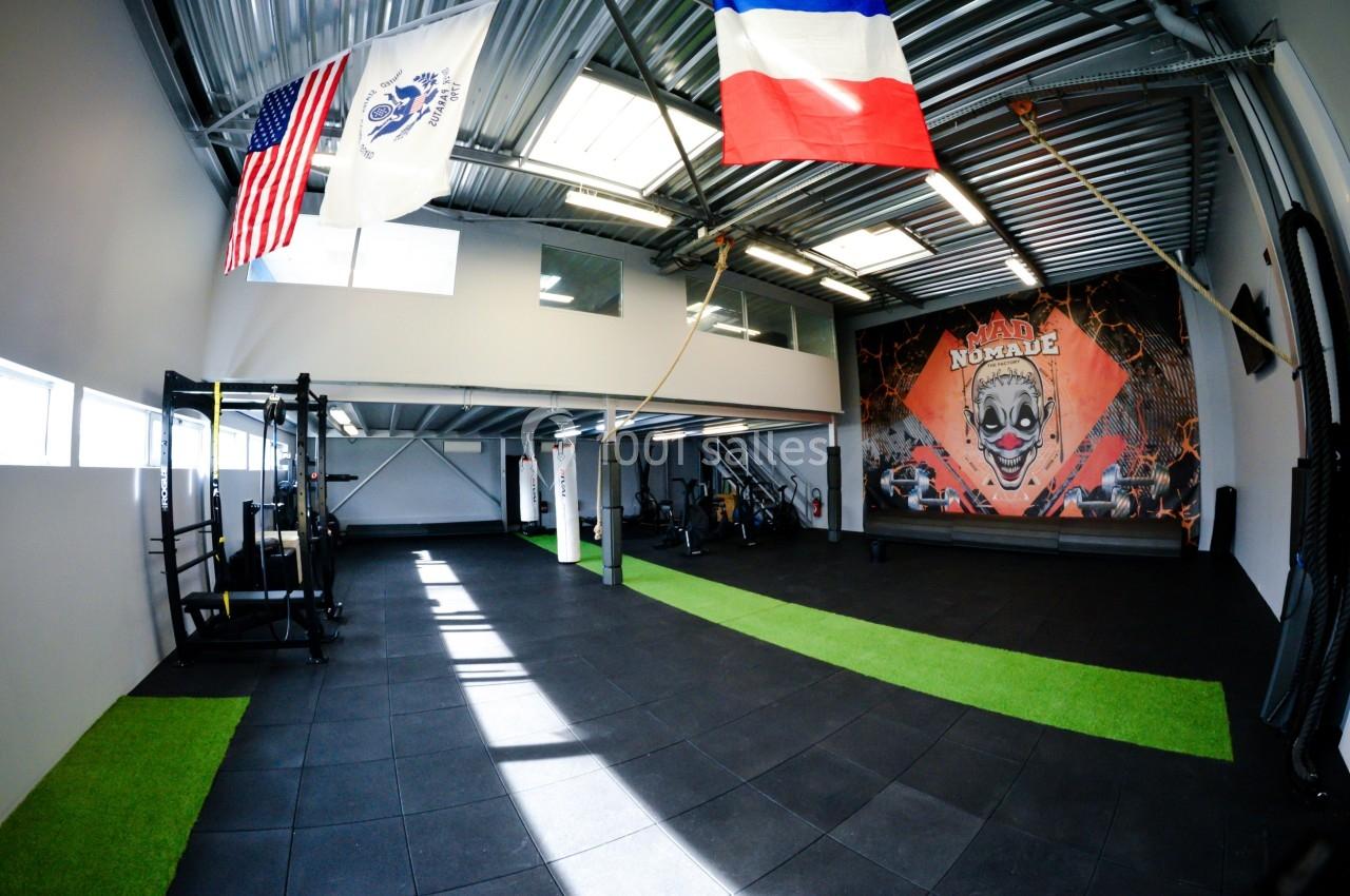 Salle de sport avec équipements, sol en dalles noires, bandes de gazon synthétique et drapeaux suspendus au plafond.