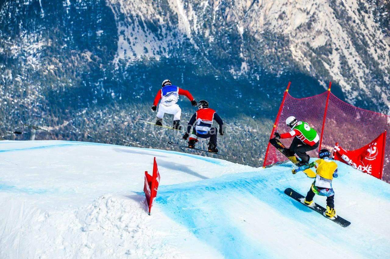 Quatre snowboardeurs en pleine course sur une piste enneigée avec des filets de sécurité rouges en arrière-plan.