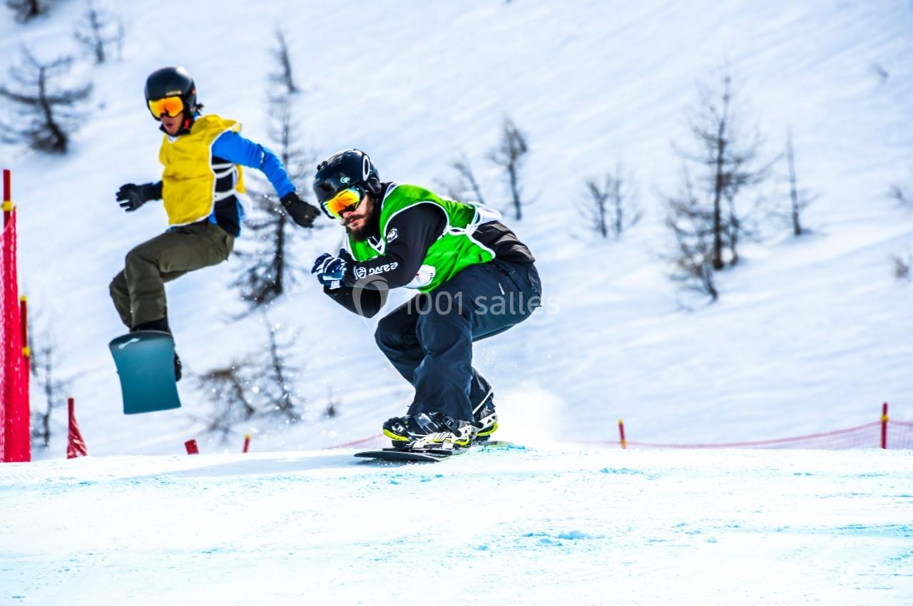 Deux snowboardeurs en pleine descente sur une piste enneigée, entourés de filets de sécurité et d'arbres.