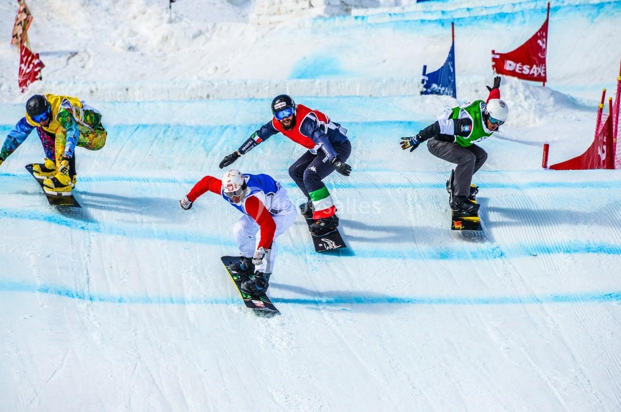 Quatre snowboardeurs en compétition descendent une piste enneigée avec des drapeaux colorés en arrière-plan.