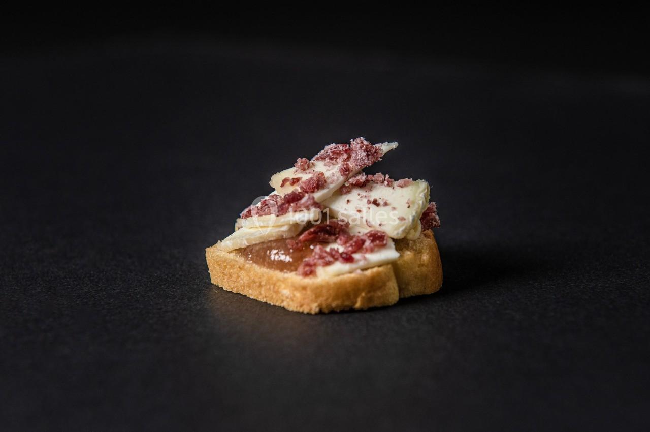 Une tranche de pain garnie de fromage, éclats de jambon et une sauce brune, posée sur une surface sombre.