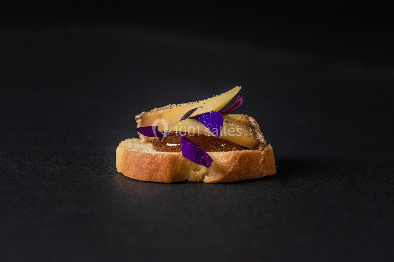 Tranche de pain garnie de fromage, fruits et pétales comestibles sur fond noir.