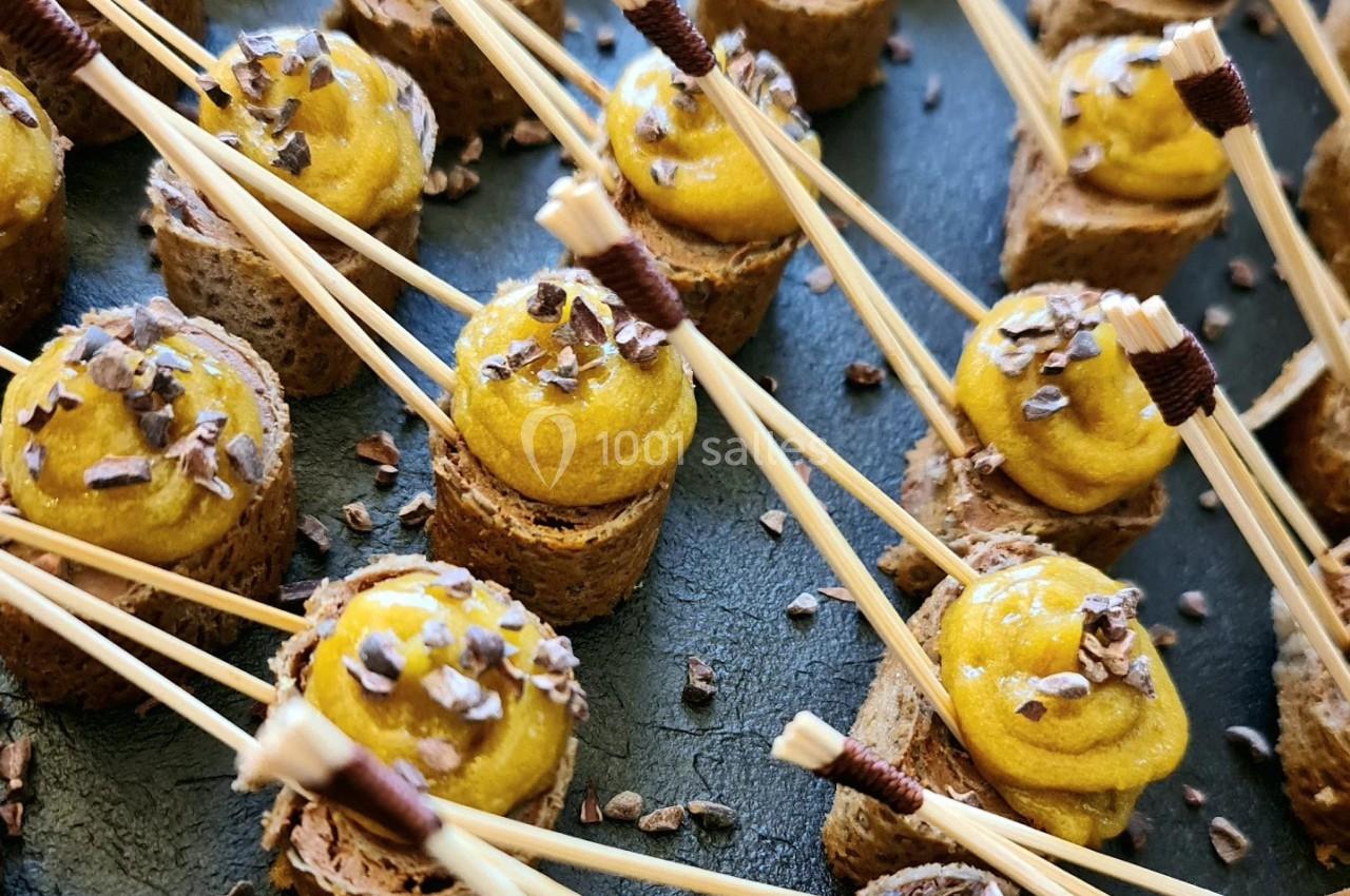 Bouchées apéritives sur pain brun, garnies de mousse jaune et décorées de copeaux, présentées avec des piques en bois.