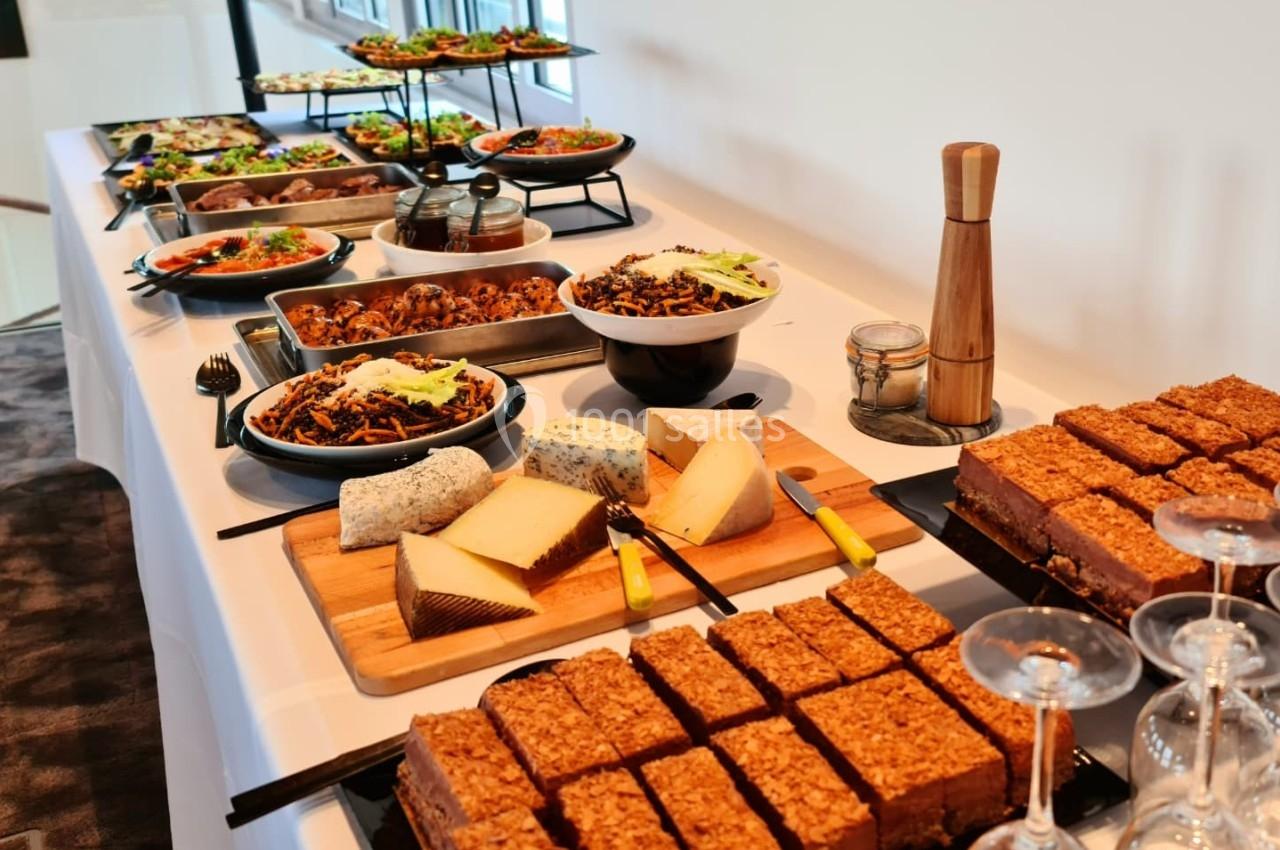 Buffet avec plats variés, fromages, desserts et couverts, disposé sur une table blanche dans un espace lumineux.