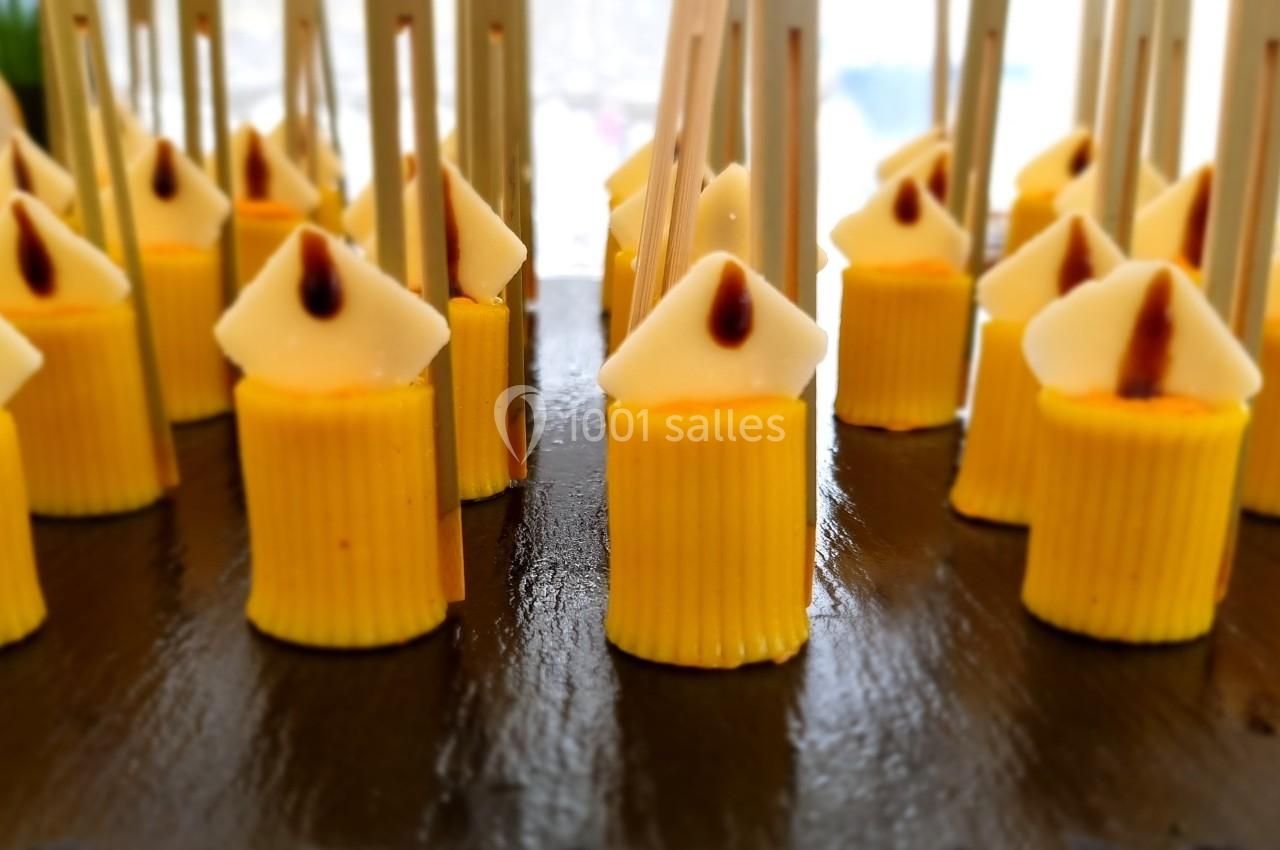 Petits desserts jaunes en forme de bougies avec des bâtonnets en bois, disposés sur une surface noire.