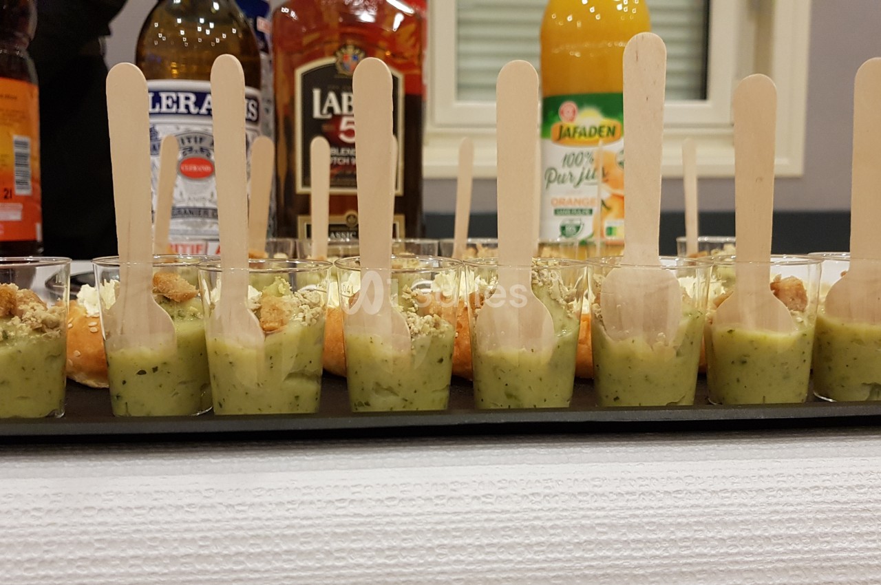 Petits verres de soupe verte avec cuillères en bois alignés sur un plateau, boissons en arrière-plan.