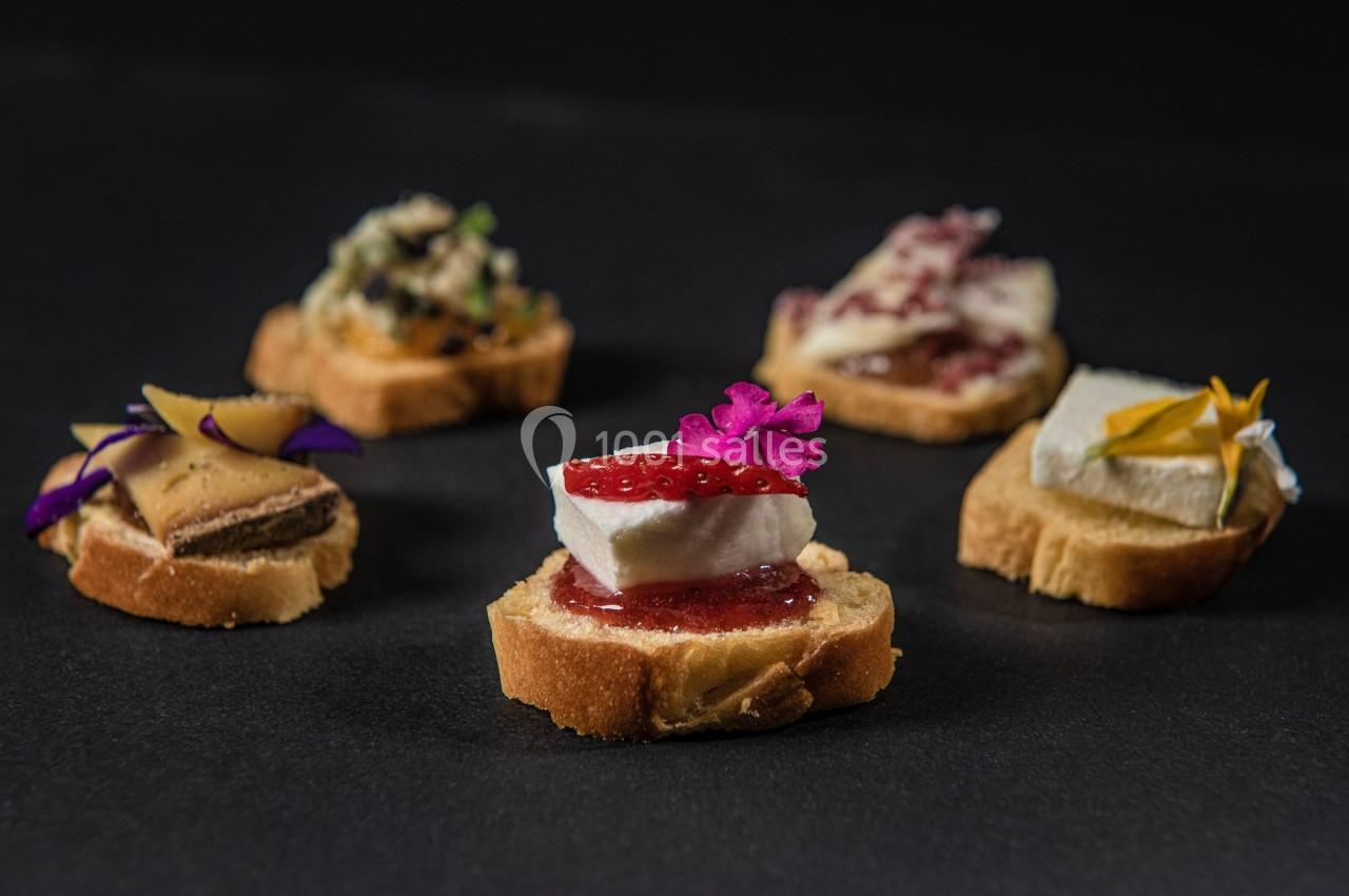 Assortiment de canapés garnis de fromage, confiture, fleurs comestibles et autres ingrédients variés sur fond noir.