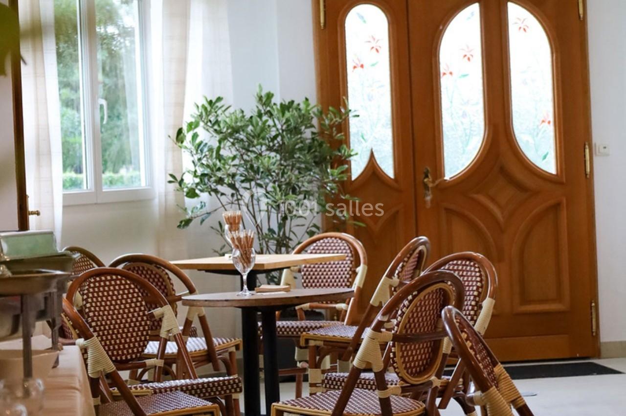 Salle lumineuse avec des chaises en rotin autour de tables, près d'une porte en bois sculpté et d'une fenêtre.