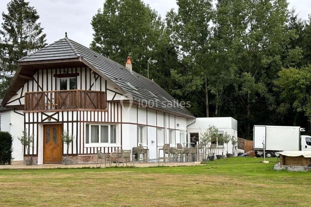 Maison à colombages avec balcon, entourée de pelouse, de tables extérieures et d'arbres en arrière-plan.