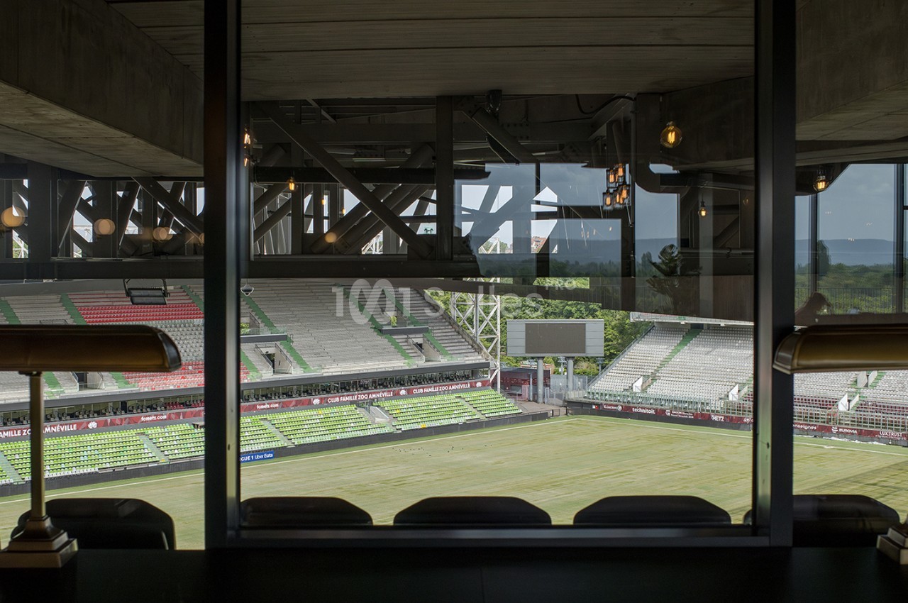 Vue depuis une loge vitrée sur un stade de football vide, avec gradins et pelouse partiellement éclairés.
