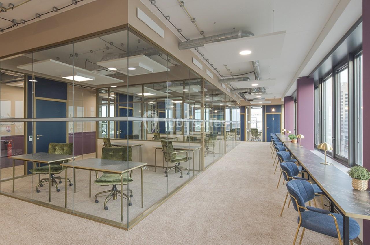 Espace de travail moderne avec bureaux vitrés, chaises vertes, et longue table avec sièges près de grandes fenêtres.