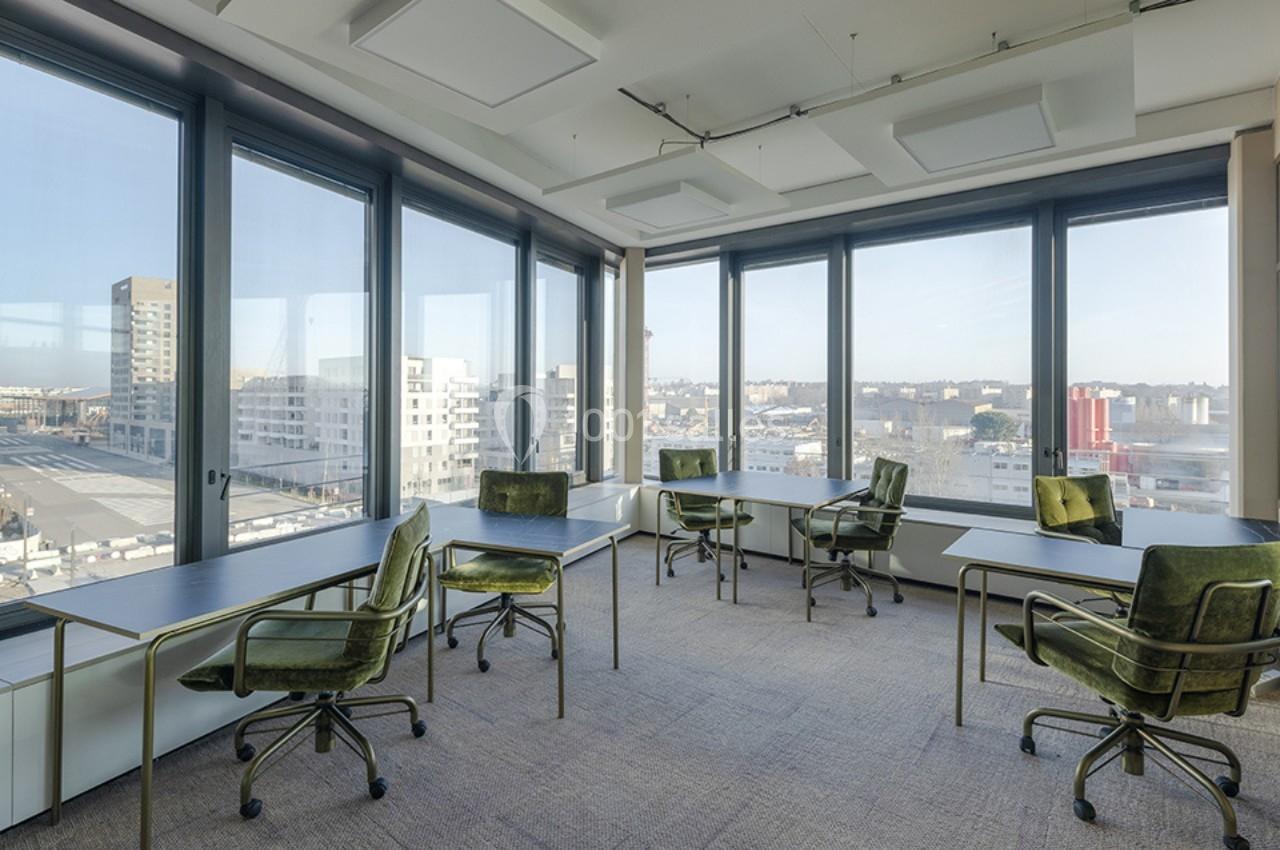 Salle de travail lumineuse avec de grandes baies vitrées, des bureaux espacés et des chaises vertes pivotantes.