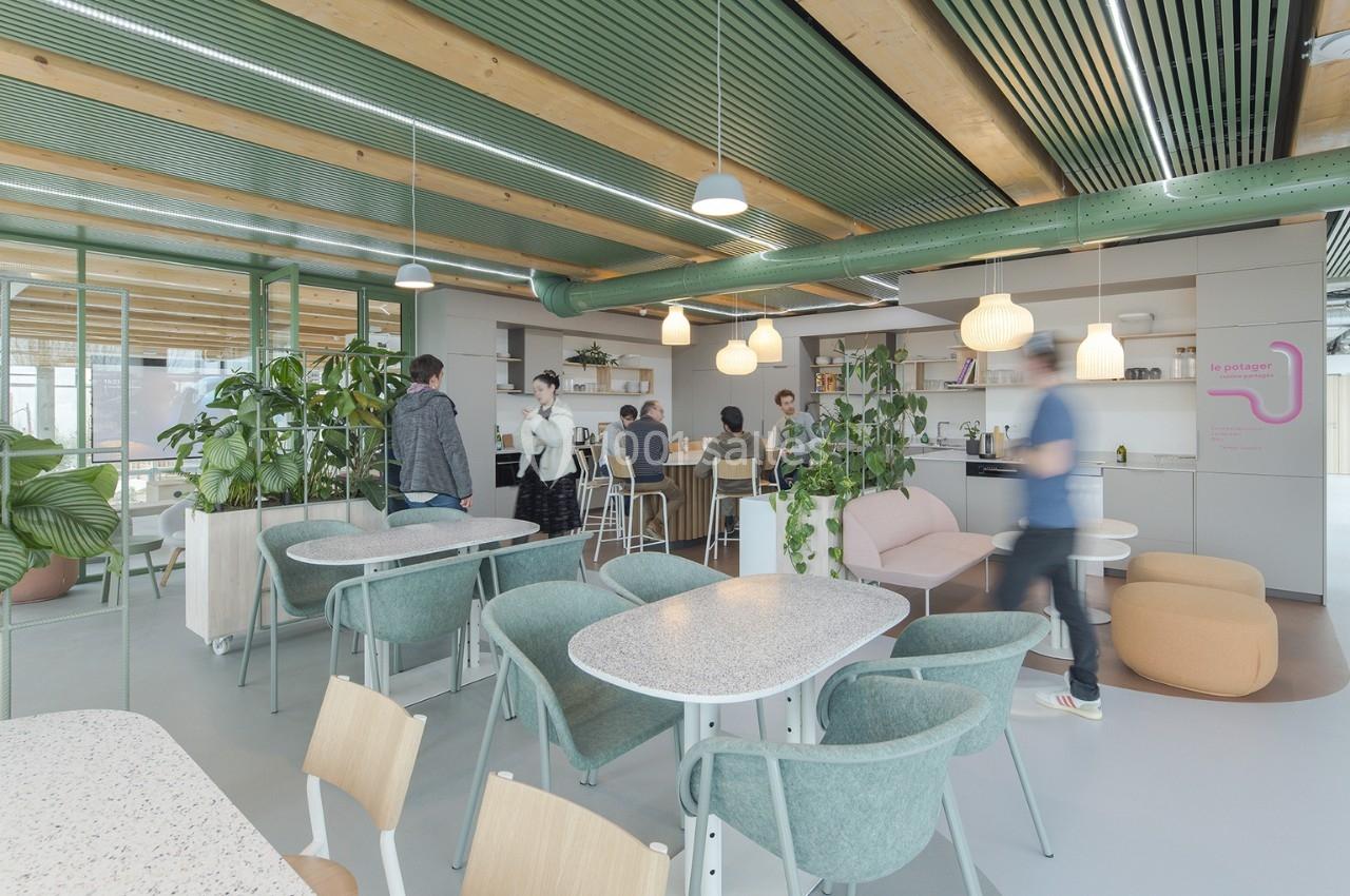 Espace de coworking lumineux avec des plantes, des tables, des chaises et des personnes discutant près d'une kitchenette.