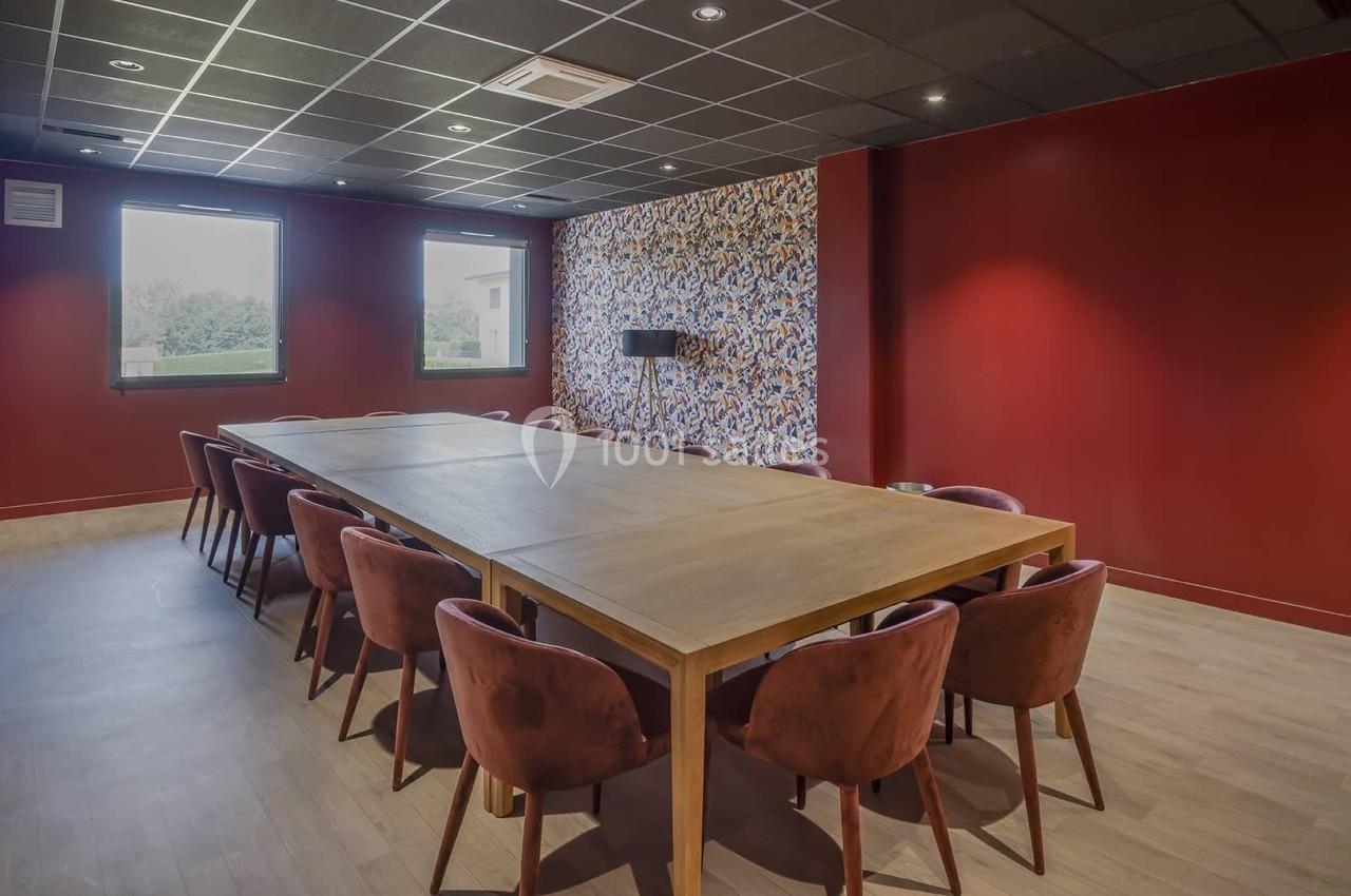 Salle de réunion avec murs rouges, grande table en bois entourée de chaises et lumière naturelle provenant des fenêtres.