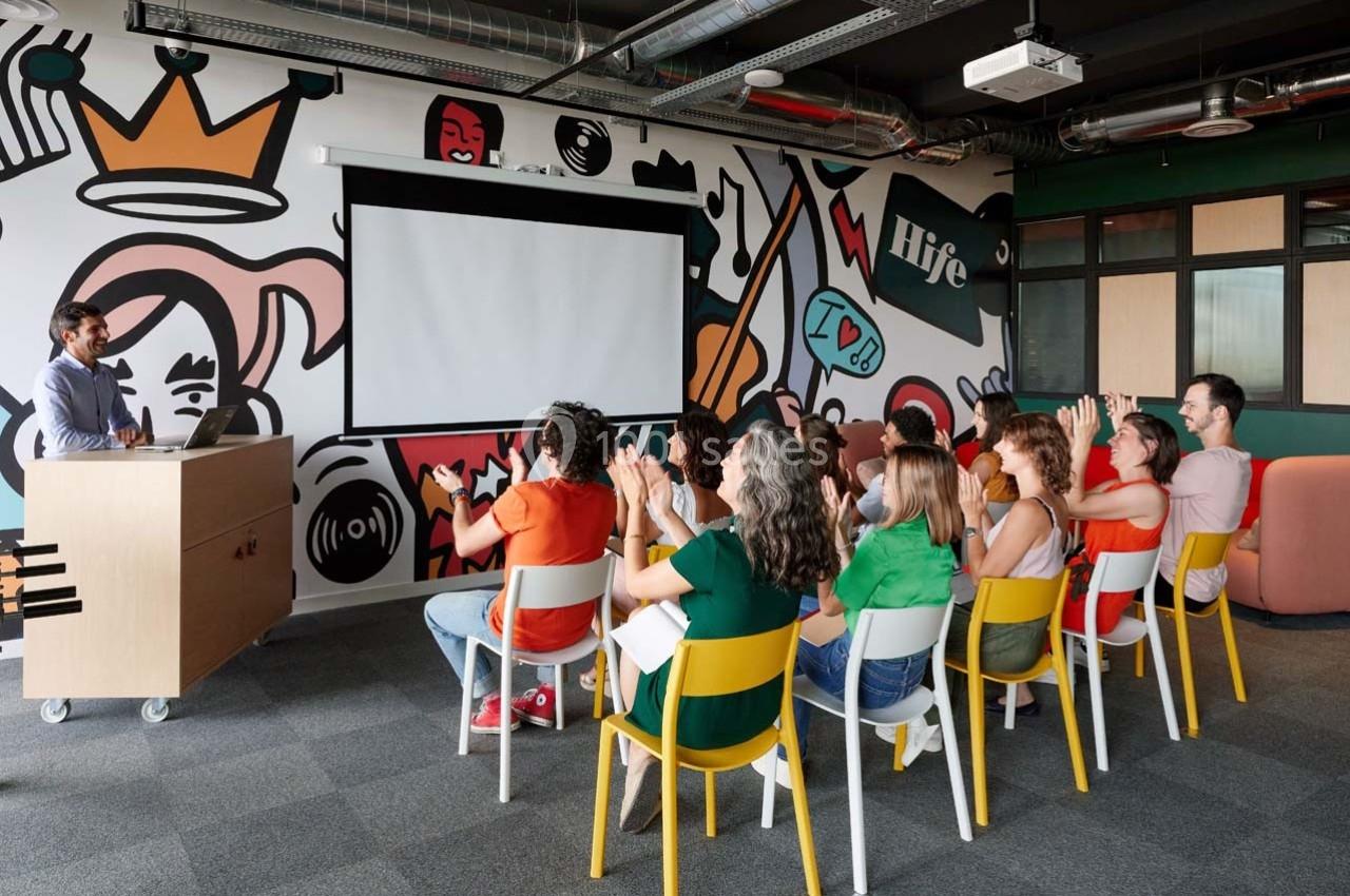 Un groupe de personnes assises dans une salle colorée, écoutant un orateur près d'un écran de projection.