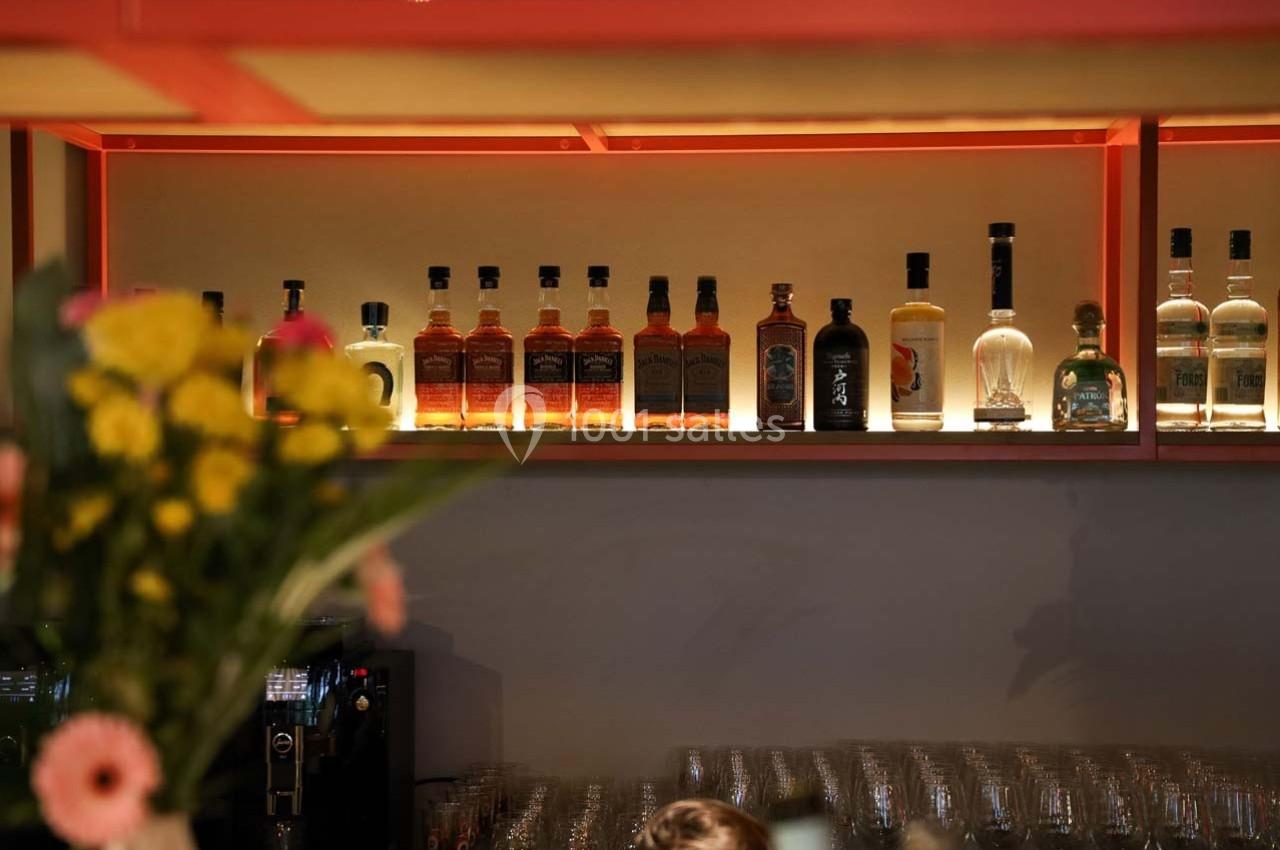 Bouteilles d'alcool alignées sur une étagère rétroéclairée, avec un bouquet de fleurs flou au premier plan.