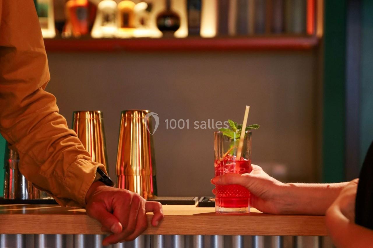 Un cocktail rouge garni de menthe est tenu par une main sur un comptoir de bar en bois, avec des shakers en arrière-plan.