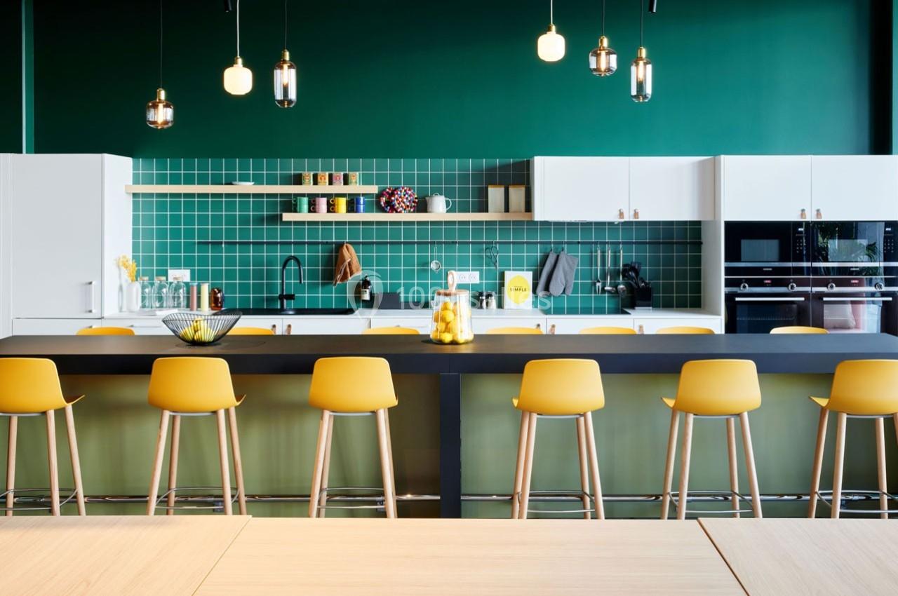 Cuisine moderne avec murs verts, îlot central noir, tabourets jaunes, étagères ouvertes et éclairage suspendu.