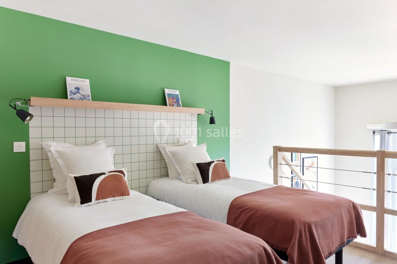Deux lits simples avec linge blanc et couvertures terracotta, dans une chambre lumineuse avec un mur vert et une rambarde en…