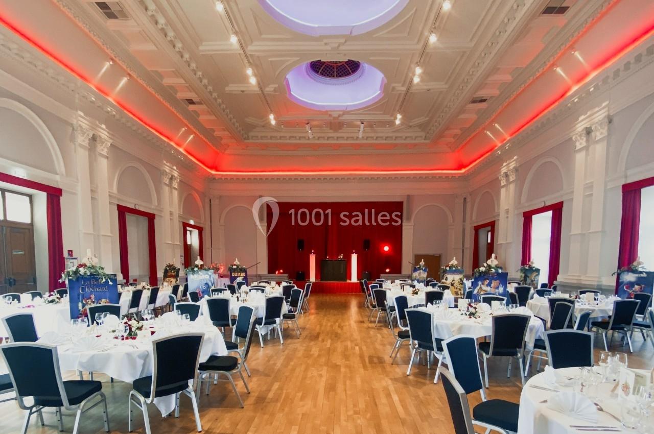 Salle de réception élégante avec tables dressées, chaises noires et rideaux rouges, éclairage tamisé au plafond.