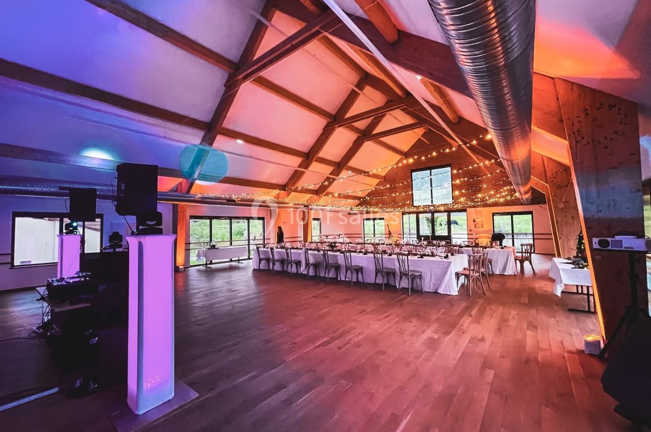 Salle de réception spacieuse avec tables dressées, éclairage chaleureux et poutres apparentes en bois.