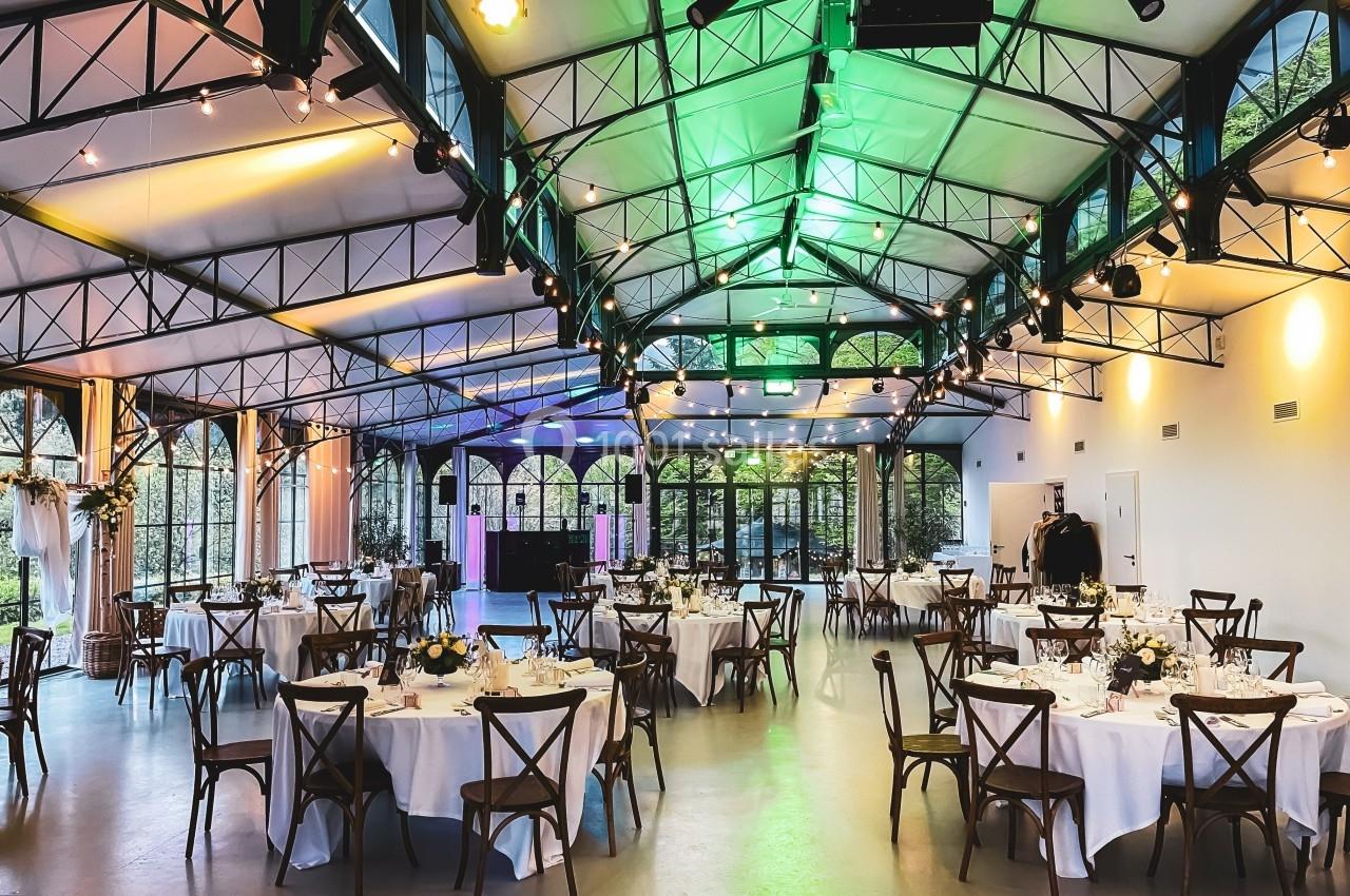 Salle de réception lumineuse avec tables rondes dressées, chaises en bois et éclairage coloré sous une verrière.