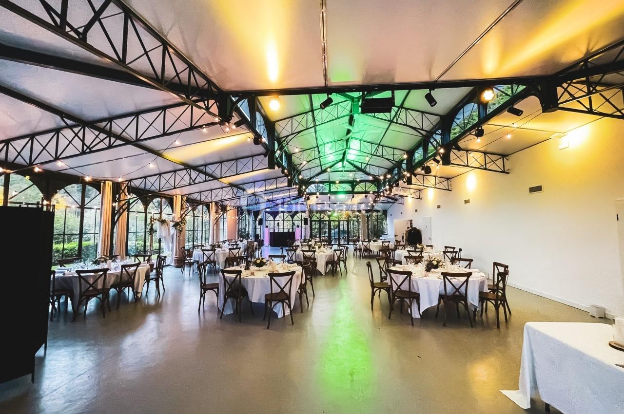 Salle de réception lumineuse avec tables rondes dressées, grandes baies vitrées et structure métallique apparente.