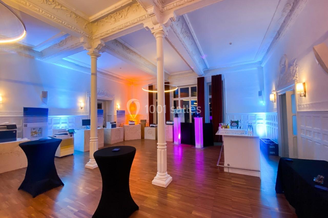 Salle élégante avec parquet, colonnes blanches, éclairage coloré et tables hautes noires.