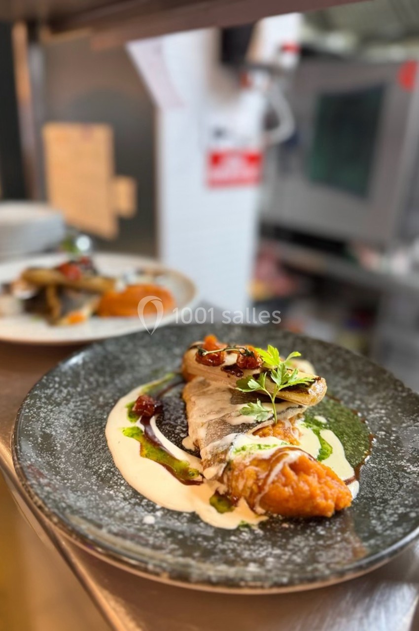 Assiette de poisson grillé avec purée, sauce crémeuse, légumes et herbes fraîches, servie dans une cuisine professionnelle.