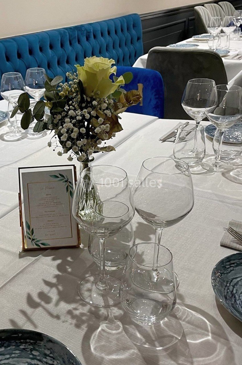 Table dressée avec nappes blanches, vaisselle élégante, verres, menu encadré et bouquet de fleurs en centre de table.