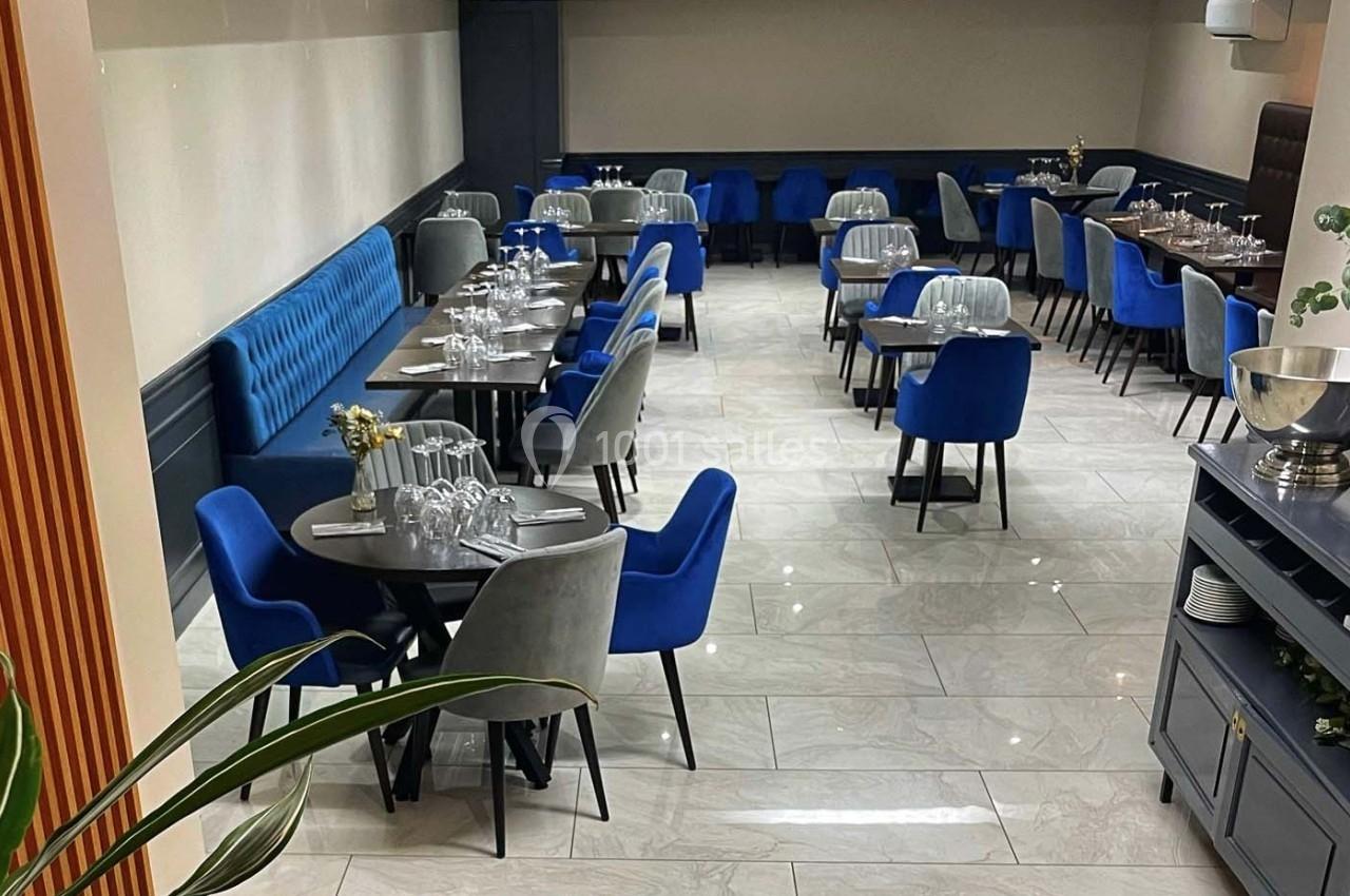 Salle de restaurant élégante avec tables dressées, chaises bleues et banquettes, sol en carrelage clair.
