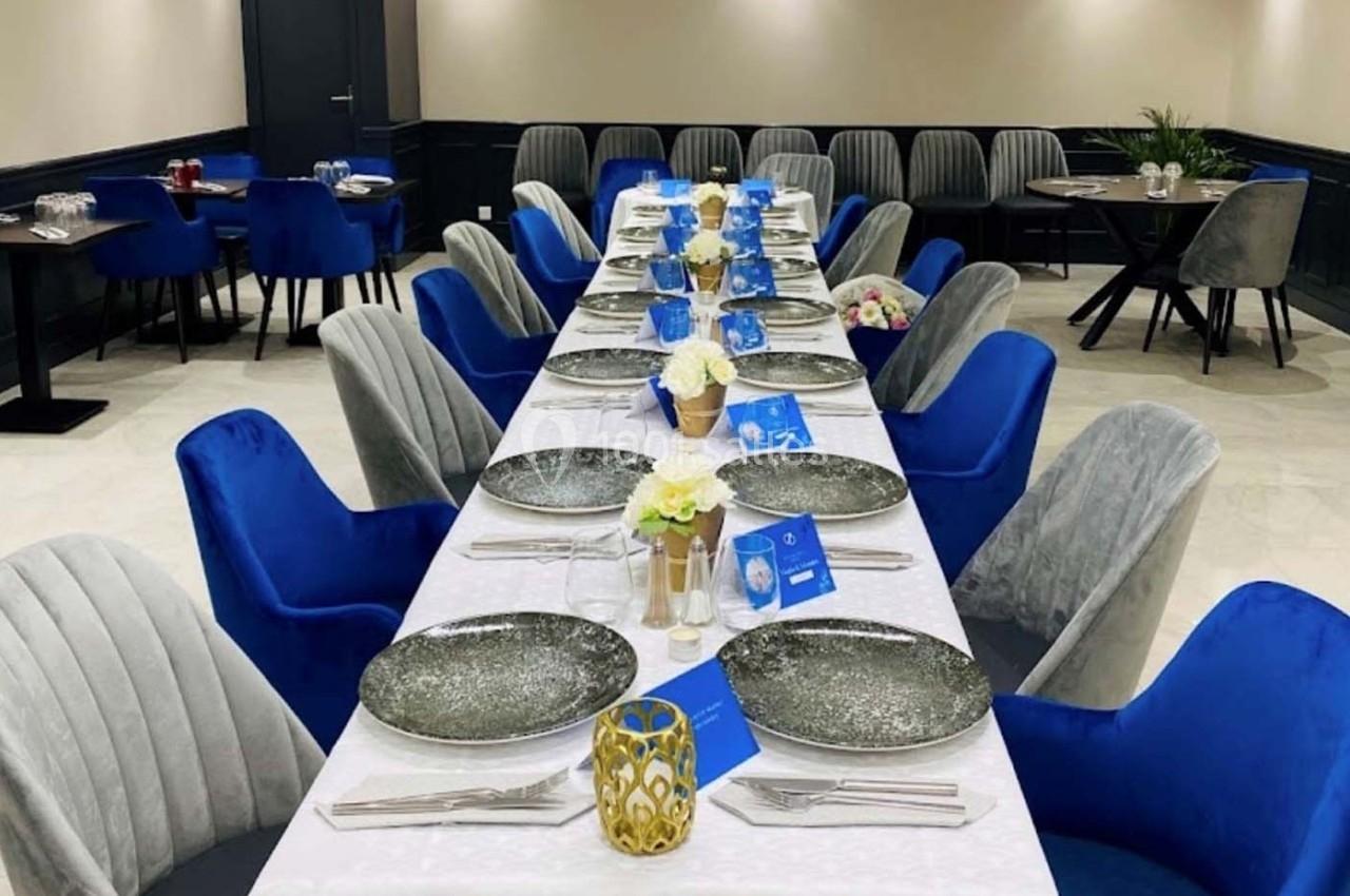 Table dressée avec nappes blanches, assiettes grises, chaises bleues et grises dans une salle de réception élégante.