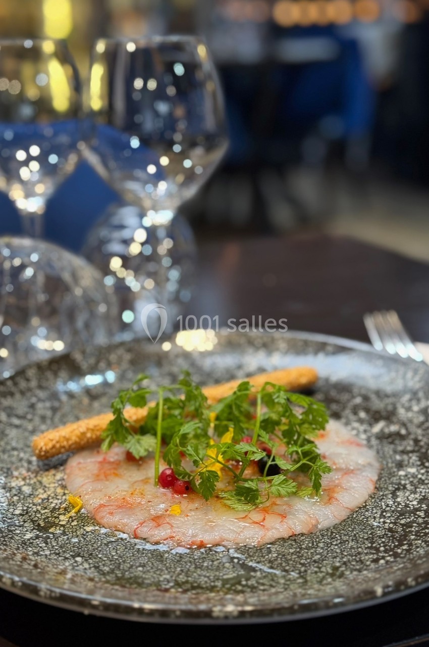 Carpaccio de crevettes décoré de fines herbes, baies rouges et accompagné d'un gressin, servi sur une assiette noire.
