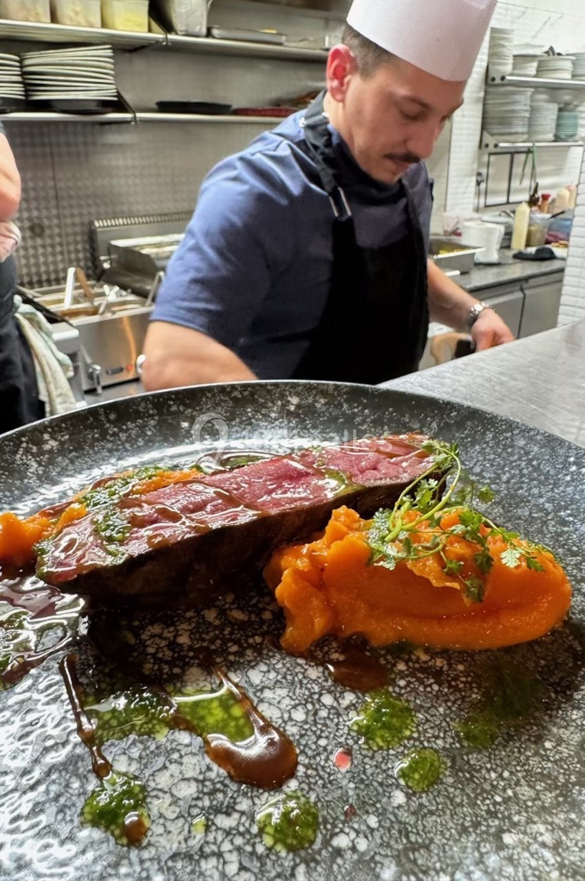 Un chef en cuisine prépare un plat de viande grillée avec purée de patate douce et sauce, présenté sur une assiette noire.