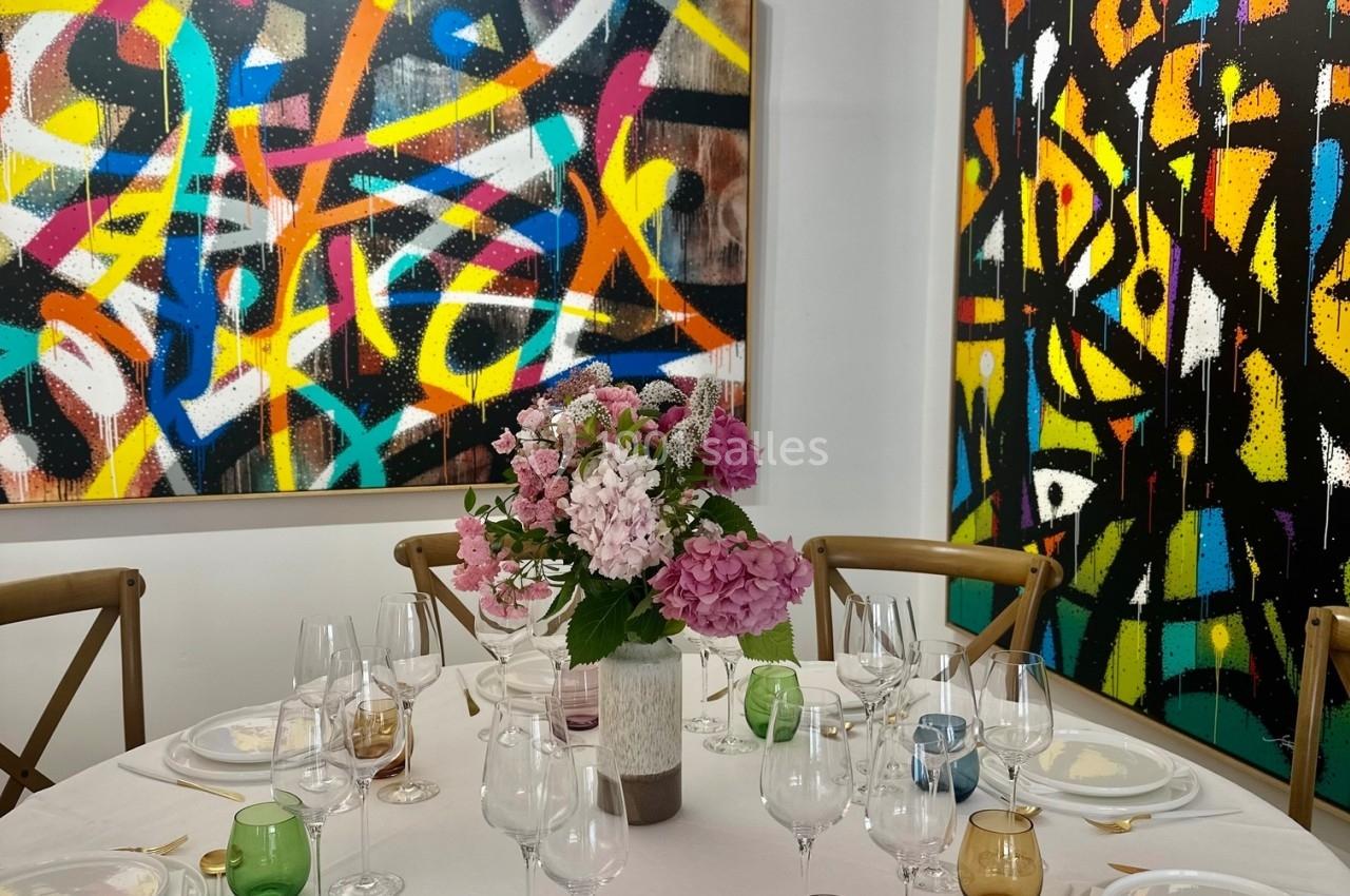 Table dressée avec vaisselle colorée et bouquet de fleurs, entourée de chaises, devant des tableaux abstraits colorés.