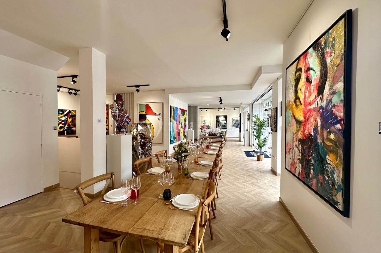Salle lumineuse avec une longue table en bois dressée, entourée de peintures colorées exposées sur les murs.