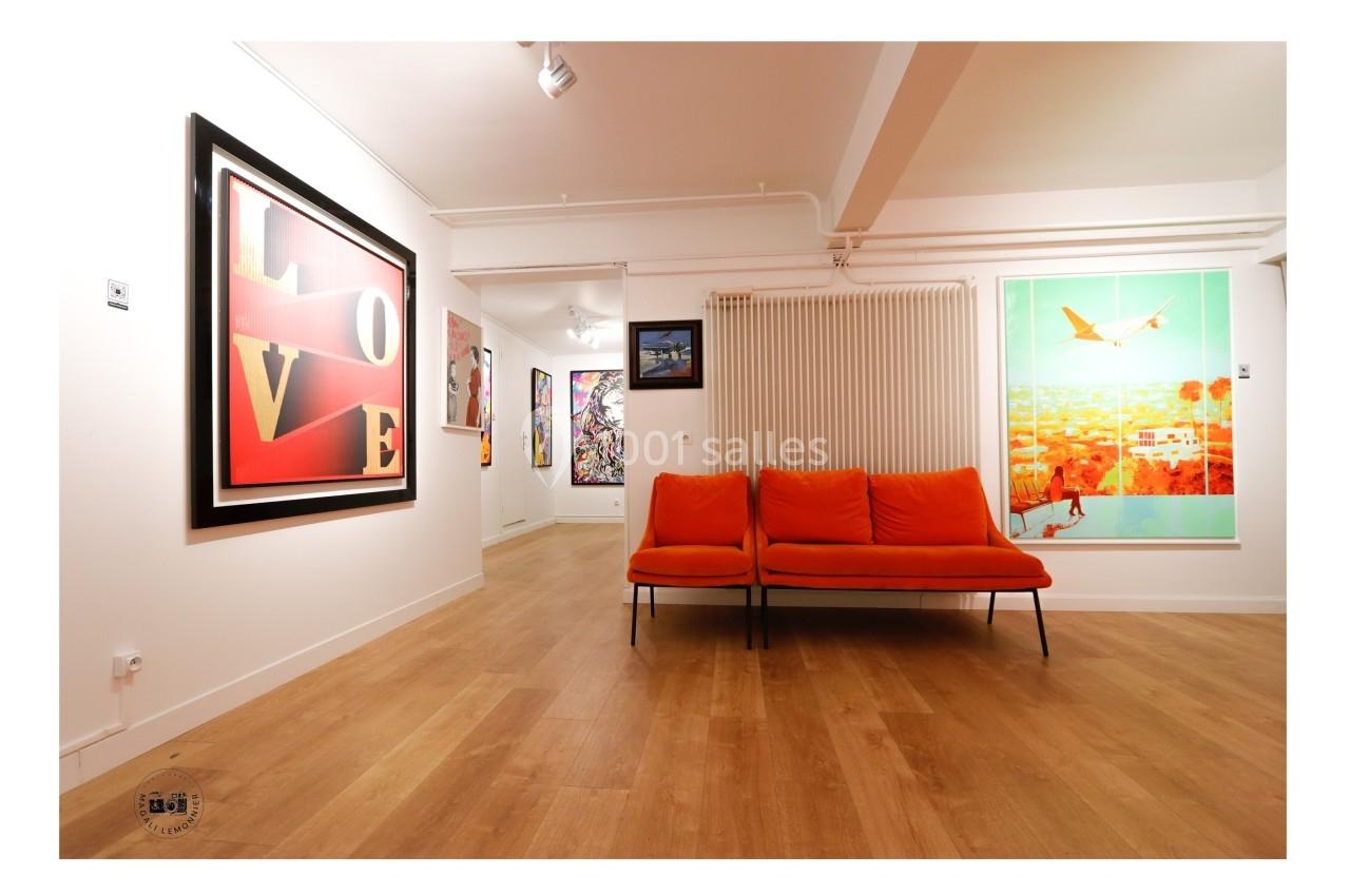 Salle d'exposition avec des œuvres d'art accrochées aux murs et un canapé orange au centre.