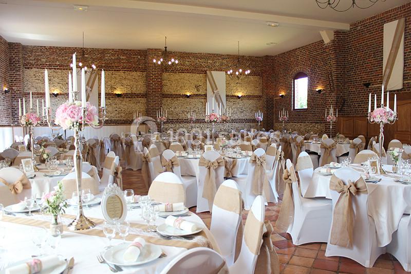 Salle de réception décorée avec des tables rondes, nappes blanches, chaises ornées de nœuds beiges et centres de table…