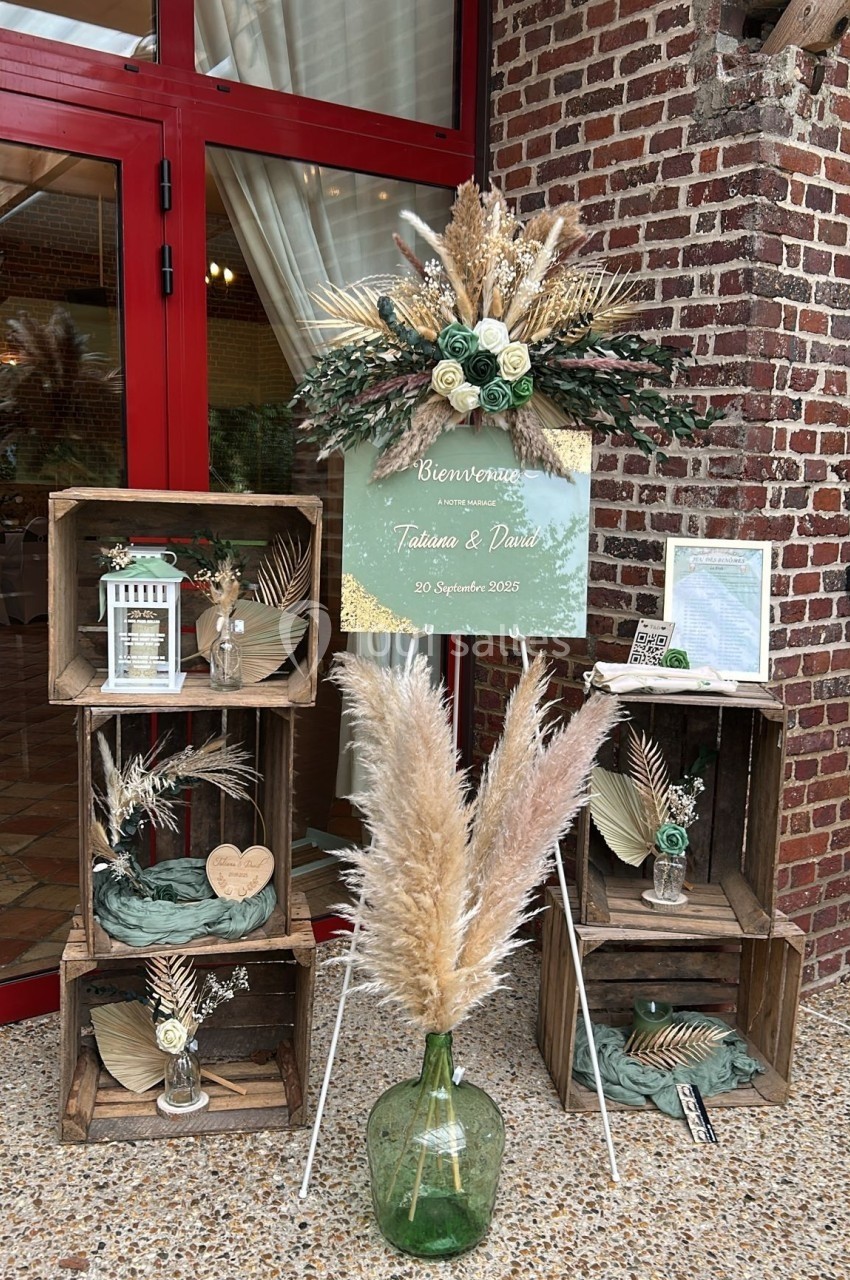 Décoration de mariage avec caisses en bois, fleurs séchées, vase en verre et panneau de bienvenue devant une porte rouge.