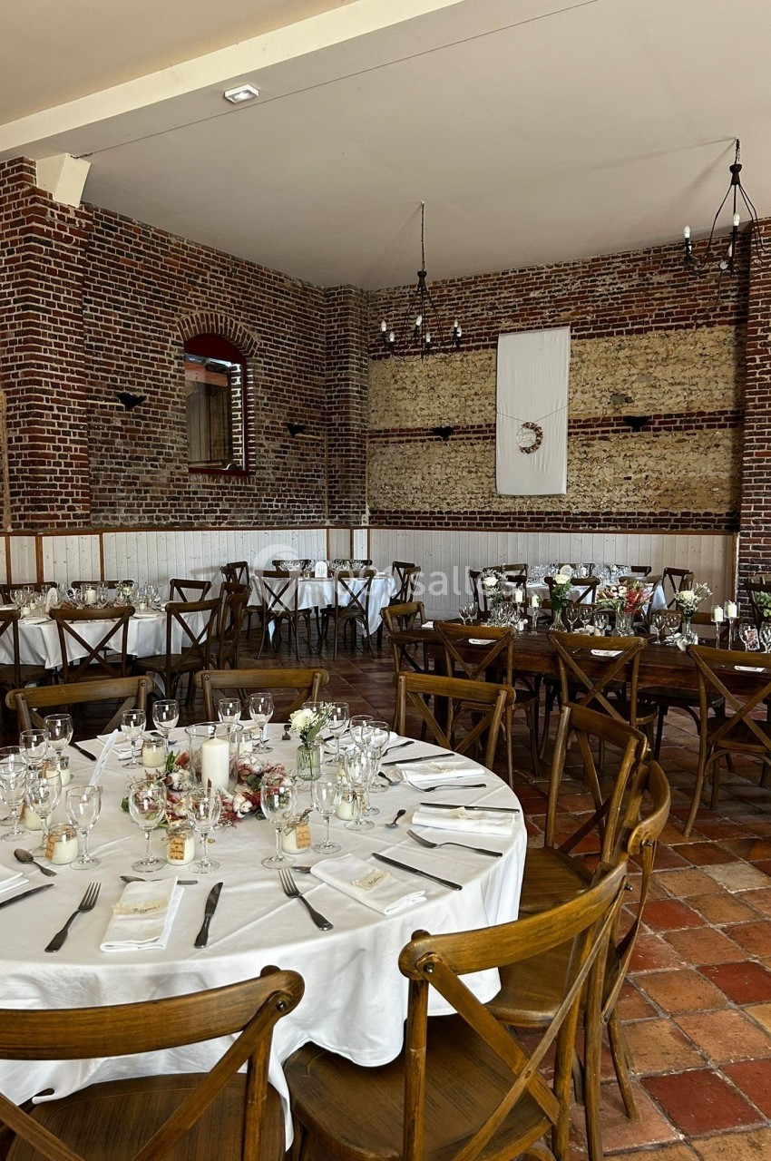 Salle de réception avec murs en briques, tables rondes dressées avec nappes blanches et chaises en bois.