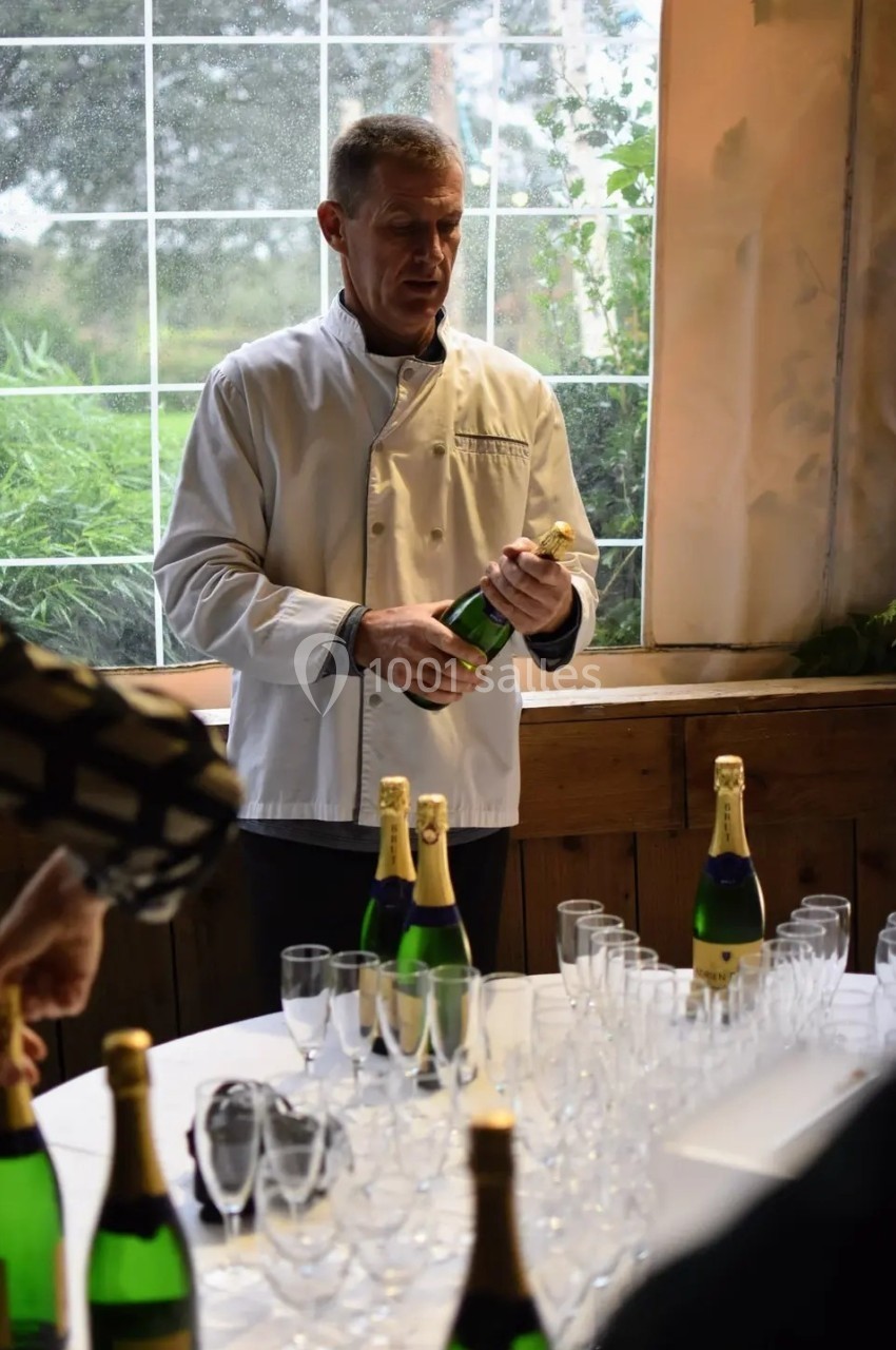 Un homme en veste de chef ouvre une bouteille de champagne devant une table garnie de verres et d'autres bouteilles.