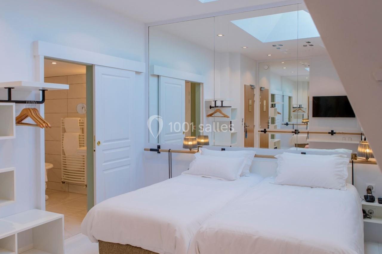 Chambre lumineuse avec lit double, miroir mural, étagères ouvertes, cintres et salle de bain attenante visible.