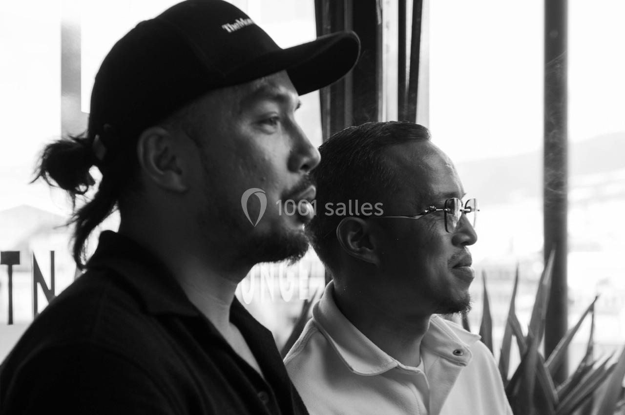 Deux hommes discutent près d'une fenêtre, l'un portant une casquette, l'autre des lunettes.