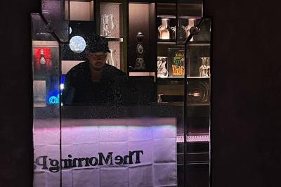 Miniature DJ Bonneville (Haute-Savoie) - Under the influence #2 Un homme portant un chapeau est derrière un comptoir, reflété dans un miroir entouré d'étagères éclairées.