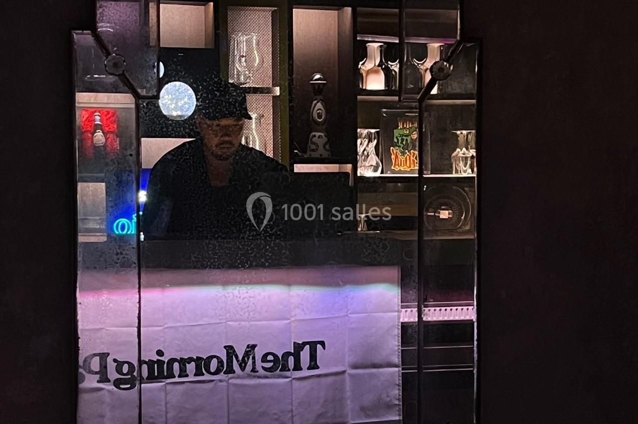 Un homme portant un chapeau est derrière un comptoir, reflété dans un miroir entouré d'étagères éclairées.