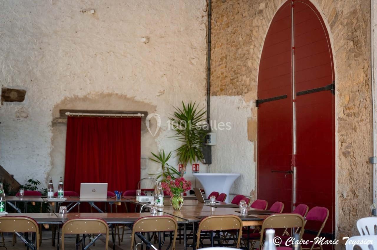 Salle de réunion avec tables disposées en U, chaises pliantes, grande porte rouge et murs en pierre apparente.